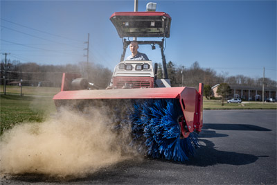 Ventrac Broom - The Ventrac KJ520 Broom effectively clears debris from a school driveway, showcasing its versatility beyond typical snow removal. With hydraulic controls for precise angling and lifting, and an optional electric actuator for adjustable broom rotation, this attachment easily handles leaves, dirt, and light gravel.