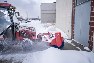 Ventrac Broom - Ventrac Broom clearing light snow from the walkway in front of a building entrance. Operating in reverse, the broom efficiently pulls snow away from the door, keeping entryways clear and accessible. Designed for precise and effective snow removal on walkways.